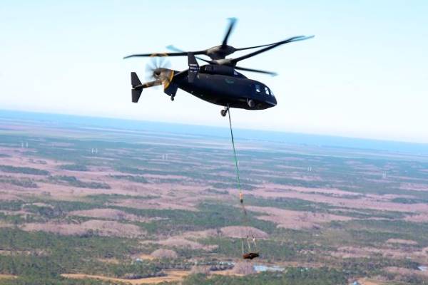 An image of the DEFIANT X helicopter in flight, carrying cargo on a cable underneath the aircraft.  